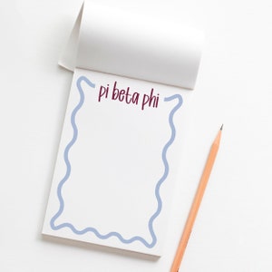 May include: A white notepad with a blue wavy border and the text "pi beta phi" written in maroon. A wooden pencil is lying on a white surface next to the notepad.