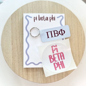 May include: A Pi Beta Phi keychain, sticker, and card set. The keychain is light blue with maroon Greek letters. The card has a wavy light blue border and the words "pi beta phi" in maroon. A round sticker with pink text is also included.