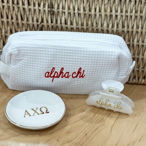 May include: White waffle-textured makeup bag with red embroidered text "alpha chi" and a white hair clip with gold text "alpha chi" and a white ceramic ring dish with gold text "AXO".