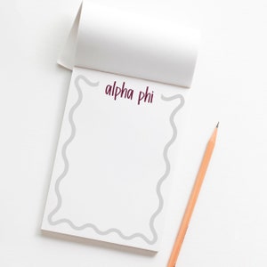 May include: A white notepad with the words "alpha phi" in burgundy script. The notepad has a gray wavy border and a wooden pencil is resting on the white surface.