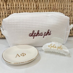 May include: White waffle-textured makeup bag with "alpha phi" embroidered in maroon. A white ceramic ring dish with "AΦ" in gold lettering. A white plastic hair clip with "alpha phi" in gold lettering.
