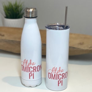 May include: Two white stainless steel tumblers with a pink and red "Alpha Omicron Pi" logo. The tumbler on the left is a water bottle with a screw-on lid. The tumbler on the right is a taller cup with a straw.