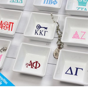 May include: White ceramic ring dishes with various sorority letters and symbols. The dishes are arranged in a grid pattern on a white wooden surface. The letters and symbols are in different colors, including pink, blue, red, and black.