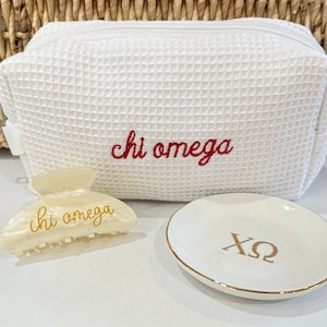 May include: White waffle-textured makeup bag with red embroidered text "chi omega". A white ceramic ring dish with gold lettering "XO" and a white plastic hair claw with gold lettering "chi omega".