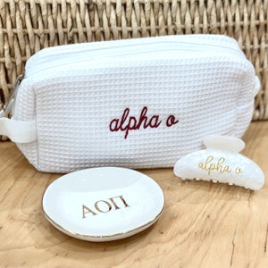 May include: White waffle-textured makeup bag with red embroidered text "alpha o". White ceramic ring dish with gold lettering "AOPI". White plastic hair clip with gold lettering "alpha o".