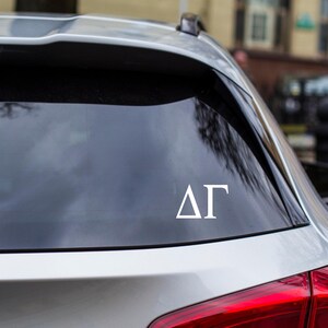 May include: White vinyl decal featuring the Greek letters Delta and Gamma (ΔΓ) on a car window. The decal is applied to the rear window of a silver car, with a blurred background.