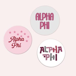 May include: Three round stickers with the text "Alpha Phi". One sticker is light pink with dark pink text and stars. Another is white with pink text. The third is white with multi-colored text.