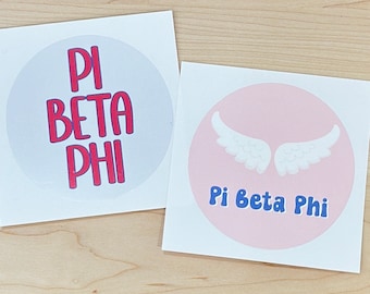 May include: Two round stickers with the text "Pi Beta Phi" in red and blue lettering. One sticker has a white background and the other has a pink background with white angel wings.