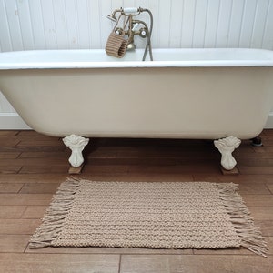 May include: A white clawfoot bathtub with a brown bath mat in front of it. The bathtub has a gold faucet and a white tiled wall behind it.