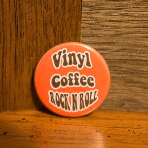 May include: A round orange button with the text "Vinyl Coffee Rock N Roll" in black.
