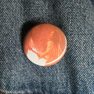 Puede incluir: Un botón redondo con una imagen de una persona tocando una guitarra frente a una puesta de sol. La persona tiene el pelo largo y castaño y lleva una camisa blanca.