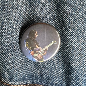 Puede incluir: Un botón redondo con una foto en blanco y negro de un músico tocando la guitarra. El músico lleva una camisa de manga larga y tiene el pelo largo.