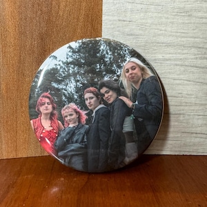 May include: A round button featuring a group of five people standing in front of a forest background. The people are wearing casual clothing and are smiling. The button is a light gray color with a white border.