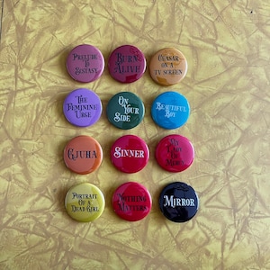 May include: Twelve round button pins with various colors and text. The text on the pins includes "Prelude to Ecstasy", "Burn Alive", "Ceasar on a TV Screen", "The Feminine Urge", "On Your Side", "Beautiful Boy", "Gjuha", "Sinner", "My Lady of Mercy", "Portrait of a Dead Girl", "Nothing Matters", and "Mirror".