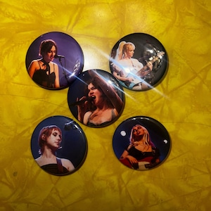 May include: Five round button pins featuring different women musicians. Each pin has a blue background with a white border. The women are wearing different outfits and are holding musical instruments.