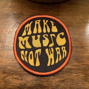 May include: A round, black patch with an orange border. The patch has yellow embroidered text that reads "MAKE MUSIC NOT WAR" in a retro font.
