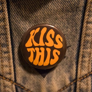 May include: A round button with the words "KISS THIS" in orange lettering on a dark brown background. The button is pinned to a denim jacket.