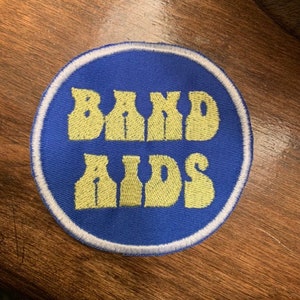 May include: A circular patch with the words "BAND AIDS" in yellow, stylized lettering. The patch has a blue background with a white border. The patch is on a wooden surface.