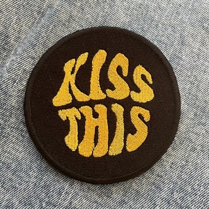 Peut inclure: Un patch rond noir avec du texte brodé jaune qui dit "Kiss This".