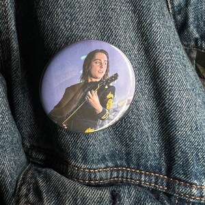 Puede incluir: Un botón redondo con una foto de una persona tocando una guitarra. La persona tiene el pelo largo y castaño y lleva una camiseta negra con un diseño amarillo. El fondo es un degradado púrpura y azul.