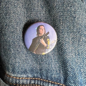 Puede incluir: Un botón redondo con fondo morado que muestra a un músico tocando la guitarra. El músico tiene el pelo largo y castaño y lleva una chaqueta negra con detalles dorados.