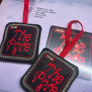 Puede incluir: Adorno de fieltro negro con texto bordado en rojo "The Pyre" y borde dorado, sujeto a una cinta roja. El adorno es de forma cuadrada y tiene la palabra "FROM" en la parte superior. El fondo incluye una funda de CD.