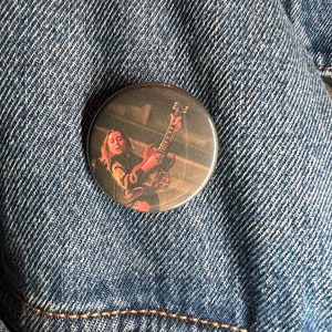 Puede incluir: Un botón redondo con una foto de una persona tocando una guitarra. La persona lleva una camisa negra y tiene el pelo largo y castaño. La guitarra es marrón y tiene una golpeador blanco. El fondo es un escenario borroso con luces.