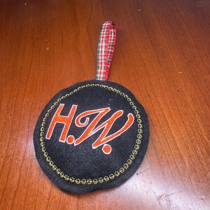 May include: A black circular ornament with the embroidered initials "H.W." in orange, outlined in white. The ornament has a gold stitched border and a red, green, and white plaid ribbon for hanging.