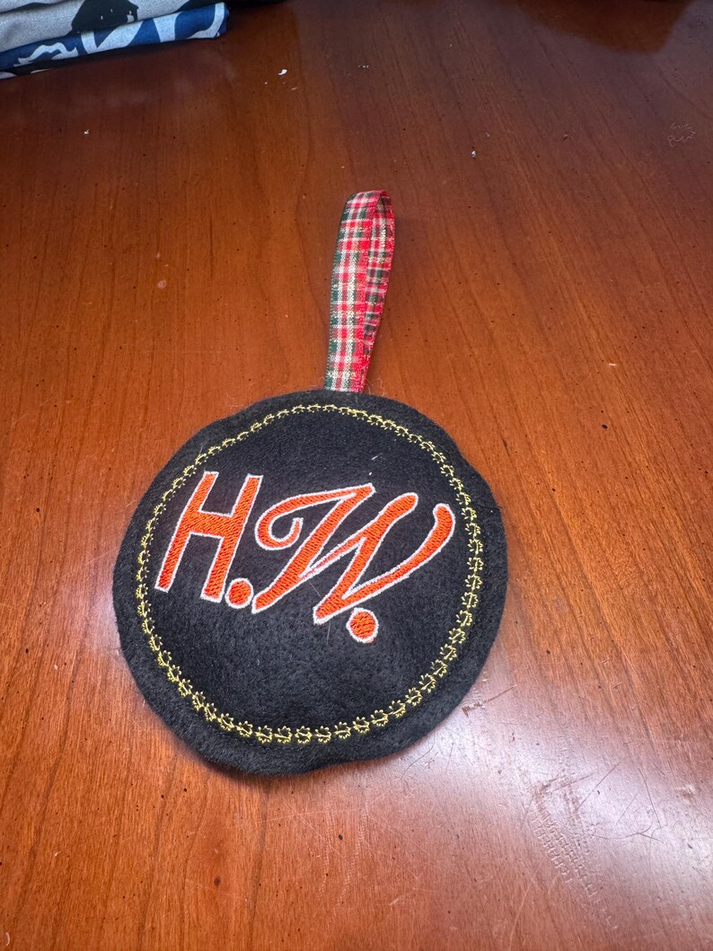 May include: A black circular ornament with the embroidered initials "H.W." in orange, outlined in white. The ornament has a gold stitched border and a red, green, and white plaid ribbon for hanging.