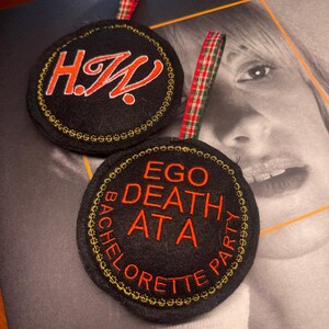 May include: Two black circular ornaments with embroidered text and gold trim. One says "H.W." in orange script, the other says "EGO DEATH AT A BACHELORETTE PARTY" in orange. Both have red and green plaid ribbon loops.