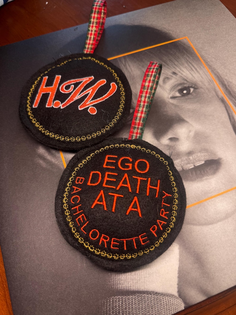 May include: Two black circular ornaments with embroidered text and gold trim. One says "H.W." in orange script, the other says "EGO DEATH AT A BACHELORETTE PARTY" in orange. Both have red and green plaid ribbon loops.