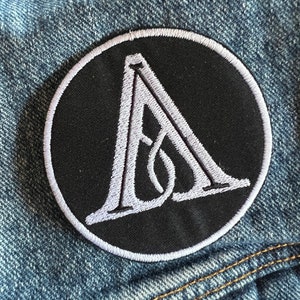 May include: A black and white embroidered patch featuring a stylised letter A within a circle. The patch is sewn onto a blue denim jacket.