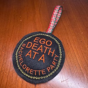 May include: Black round coaster with orange embroidered text that reads "EGO DEATH AT A BACHELORETTE PARTY". The coaster has a gold stitched border and a plaid ribbon loop.