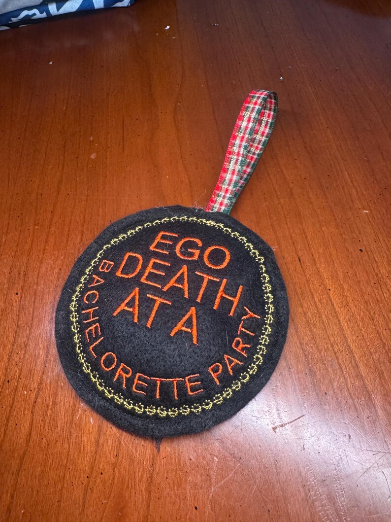 May include: Black round coaster with orange embroidered text that reads "EGO DEATH AT A BACHELORETTE PARTY". The coaster has a gold stitched border and a plaid ribbon loop.