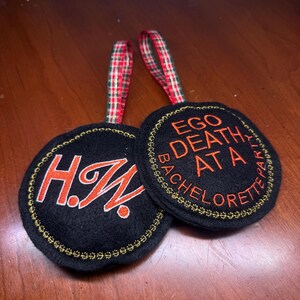 May include: Two black circular ornaments with gold embroidered edges and red and white plaid ribbon hangers. One ornament has the initials "H.W." in orange. The other says "EGO DEATH AT A BACHELORETTE PARTY" in orange.