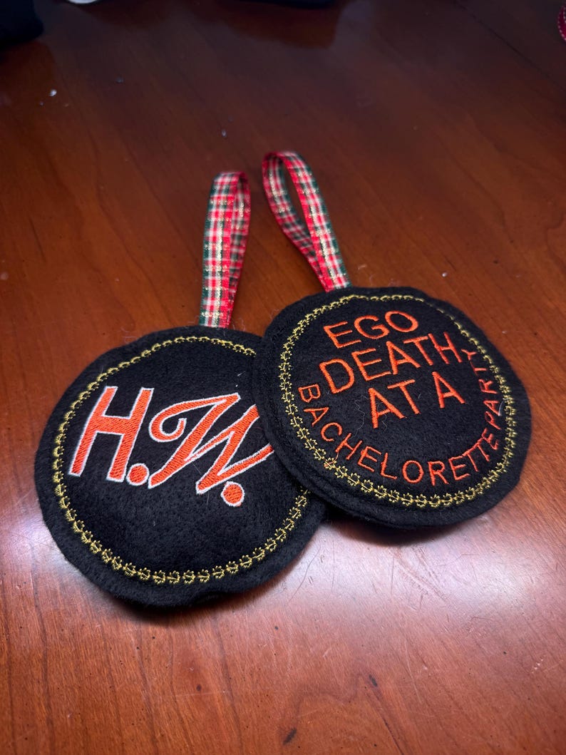 May include: Two black circular ornaments with gold embroidered edges and red and white plaid ribbon hangers. One ornament has the initials "H.W." in orange. The other says "EGO DEATH AT A BACHELORETTE PARTY" in orange.