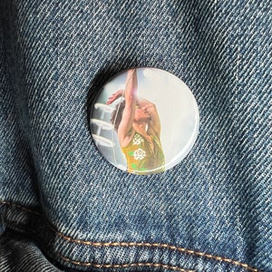 Puede incluir: Un botón blanco con una foto de una persona que lleva una camisa floral amarilla y blanca, con el brazo levantado en el aire.