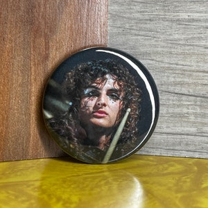 Puede incluir: Un botón redondo con una foto en blanco y negro de una persona con el pelo largo y rizado tocando la batería. La persona lleva ropa negra y tiene maquillaje oscuro en los ojos.