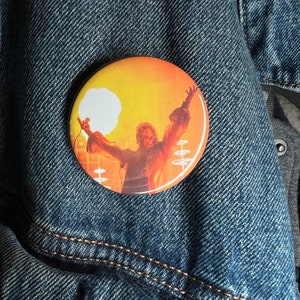 Puede incluir: Un botón redondo con una imagen de un músico tocando la batería frente a un fondo naranja y amarillo brillante.