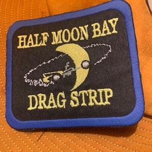 May include: A black embroidered patch with a blue border. The patch features a yellow crescent moon with a car driving around it. The text "HALF MOON BAY DRAG STRIP" is embroidered in yellow.