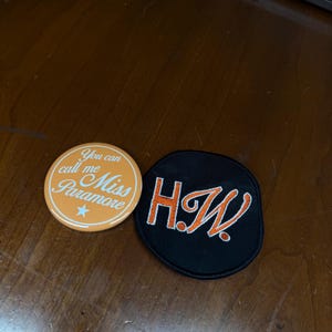 Puede incluir: Dos parches, uno naranja y otro negro. El parche naranja tiene el texto "You can call me Miss Paramore" en blanco. El parche negro tiene el texto naranja "H.W."
