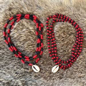May include: Two beaded necklaces with alternating red and black beads. Each necklace has a white cowrie shell pendant. The necklaces are displayed on a textured, brown and tan surface.