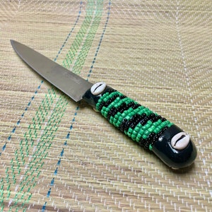 May include: A kitchen knife with a black handle and a green and black beaded design. The handle is decorated with two white cowrie shells.