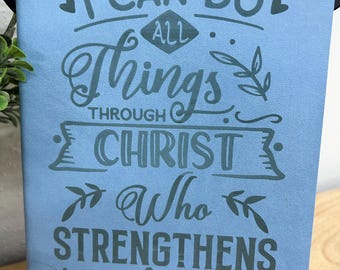 May include: A light blue journal with the inscription "I CAN DO ALL THINGS THROUGH CHRIST Who STRENGTHENS me" in a darker blue. The text is surrounded by decorative elements like hearts and leaves. The journal is displayed on a wooden surface.