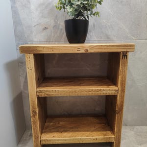 May include: A rustic wooden shelf unit with two shelves. The shelf is made of reclaimed wood and has a natural finish. A small potted plant sits on top of the shelf.