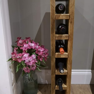 May include: A rustic wooden wine rack with six slots for bottles. The rack is made of reclaimed wood and has a natural finish. It is standing on a wooden floor with a vase of pink flowers in the corner.