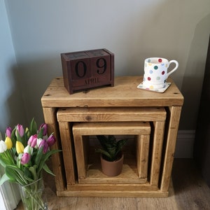 May include: Three wooden nesting tables with a natural finish. The top table has a wooden calendar block that reads "09 APRIL" and a white mug with colourful polka dots. A small potted plant sits on the bottom table.