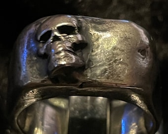 May include: A silver skull ring with a textured finish. The skull is detailed and has a three-dimensional appearance.