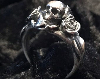 Puede incluir: Un anillo de plata con una calavera y rosas. La calavera está detallada con cuencas de los ojos y una nariz. Las rosas son estilizadas y tienen un acabado texturizado.