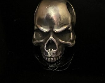 Könnte beinhalten: Ein silberner Totenkopf-Anhänger mit detailliertem Design. Der Schädel hat oben ein Loch zum Anbringen an einer Halskette oder einem Armband.